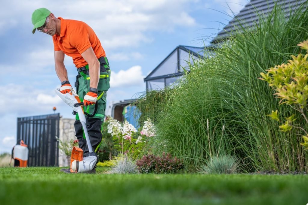 A man wearing an orange shirt is using a lawn mower to trim the grass in a well-maintained garden.